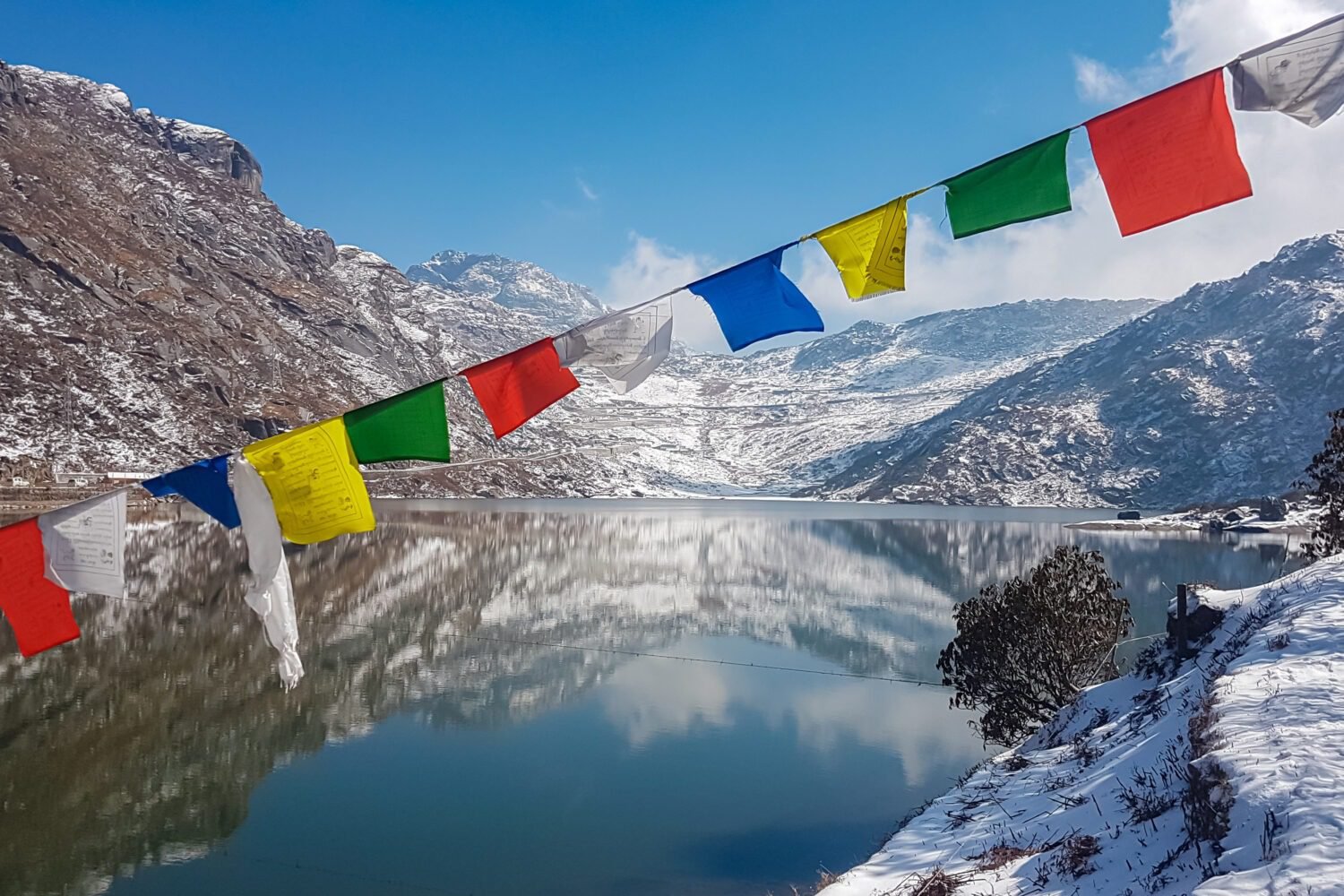 String of colorful Tibetan prayer flags at Glacial Tsomgo Lake surrounded by snow-capped mountain peaks near Gangtok, Sikkim, India. Calm water reflects sky and Himalaya landscape like mirror.