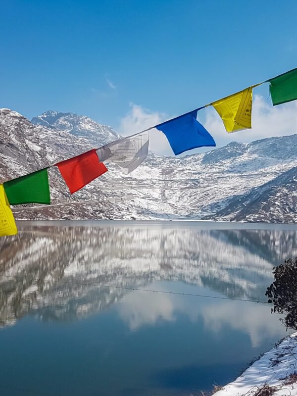 String of colorful Tibetan prayer flags at Glacial Tsomgo Lake surrounded by snow-capped mountain peaks near Gangtok, Sikkim, India. Calm water reflects sky and Himalaya landscape like mirror.