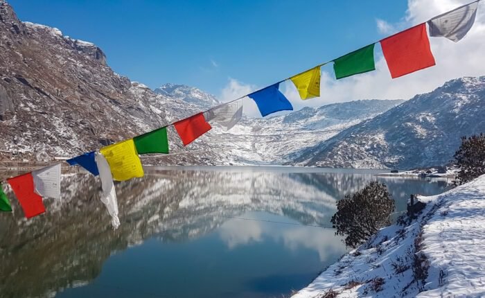 String of colorful Tibetan prayer flags at Glacial Tsomgo Lake surrounded by snow-capped mountain peaks near Gangtok, Sikkim, India. Calm water reflects sky and Himalaya landscape like mirror.