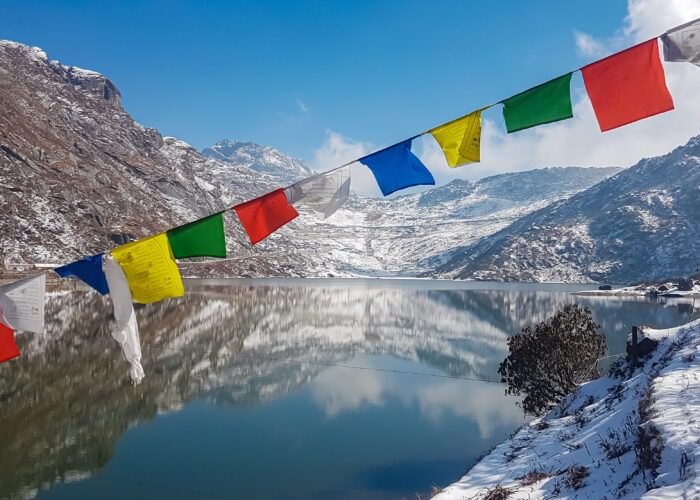 String of colorful Tibetan prayer flags at Glacial Tsomgo Lake surrounded by snow-capped mountain peaks near Gangtok, Sikkim, India. Calm water reflects sky and Himalaya landscape like mirror.